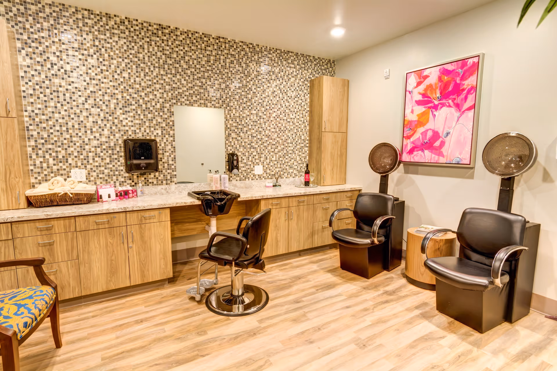 Interior view of a salon area with wooden cabinets and a mosaic tile backsplash. There is a black salon chair in front of a sink, two black chairs with hair dryers attached, a small round wooden table, and a colorful pink floral painting on the wall. The floor is light wood, and there is a patterned chair partially visible on the left.