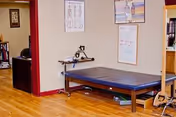 Therapy room with a treatment table against a wall, medical charts, a small desk, and hardwood floors.