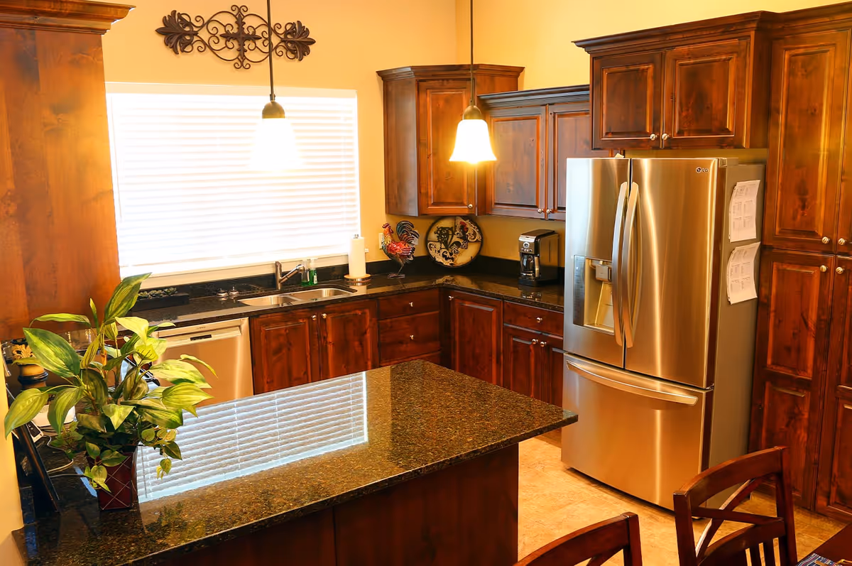A warm kitchen interior featuring dark wooden cabinets, a stainless steel refrigerator with papers attached, a double sink under a window with blinds, a coffee maker on the counter, two pendant lights hanging from the ceiling, and a granite countertop island with a potted plant.