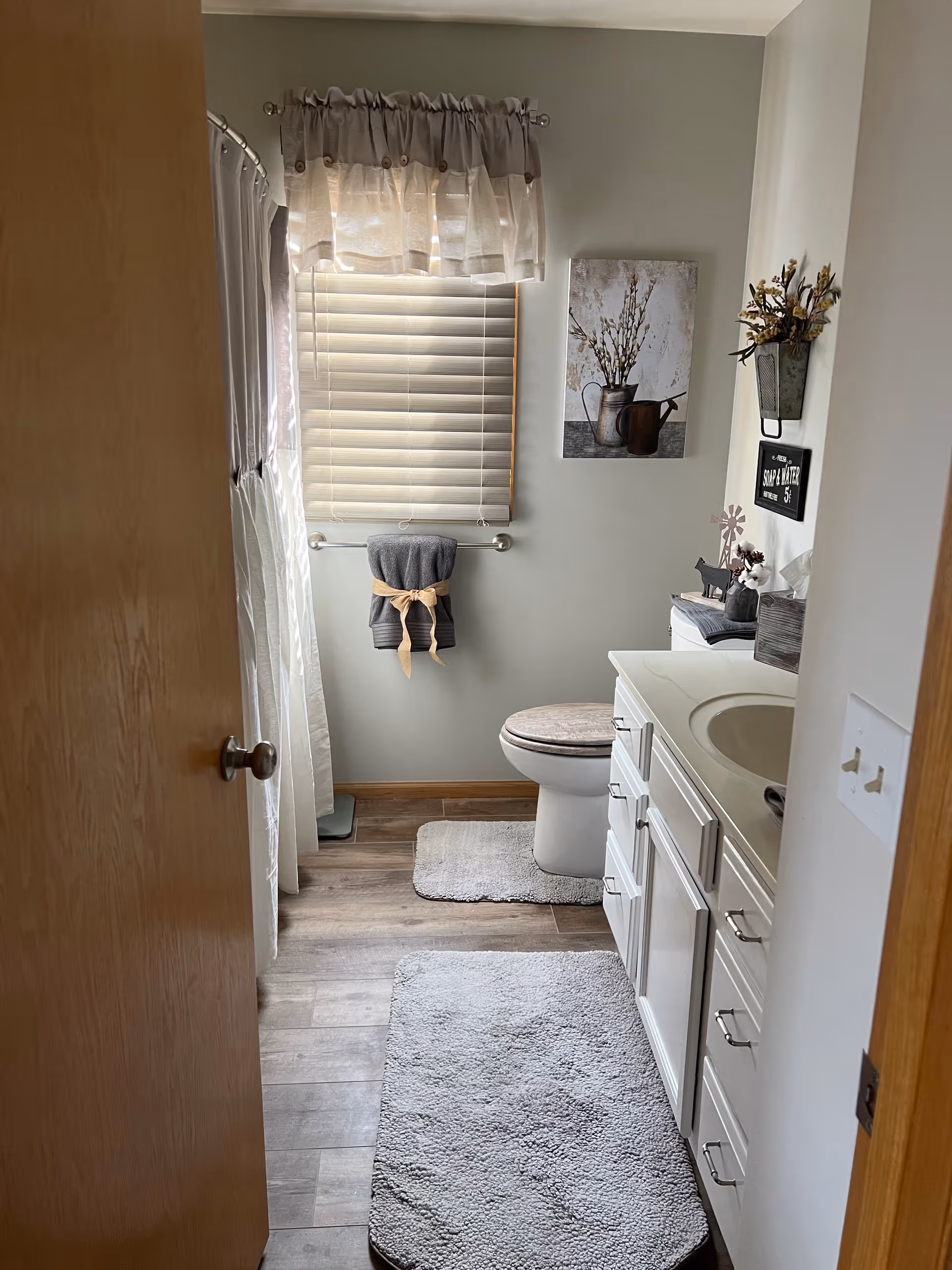 A small bathroom with a wooden door partially open. Inside, there is a white vanity with a sink on the right, a toilet with a cushioned seat cover, and a window with blinds and a sheer curtain above a towel rack holding a gray towel tied with a beige ribbon. The floor has wood-like tiles with two gray bath mats. The walls are light gray with decorative items including a framed picture of a watering can with flowers and a small wall-mounted planter with dried flowers.