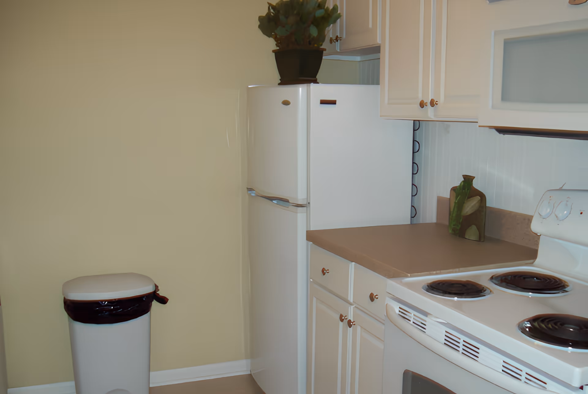 Small kitchen corner with a white refrigerator, stove, cabinets, countertop, and a trash can.