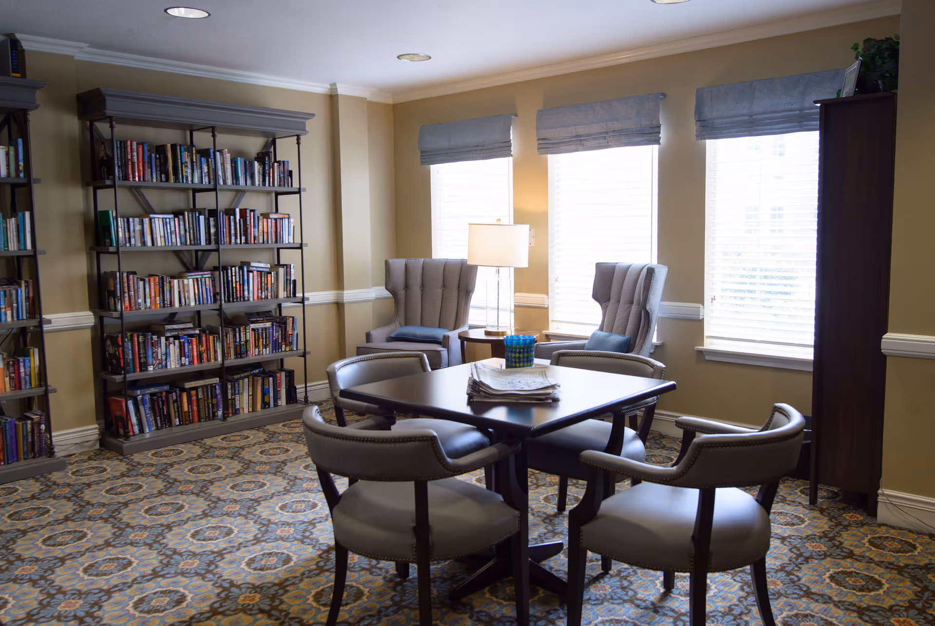 A cozy reading and sitting area with a square table surrounded by four cushioned chairs in the center. Behind the table are two wingback armchairs with blue cushions, a small round side table with a lamp, and three large windows with blinds and blue valances. To the left, there are two tall bookshelves filled with books. The room has beige walls and a patterned carpet.