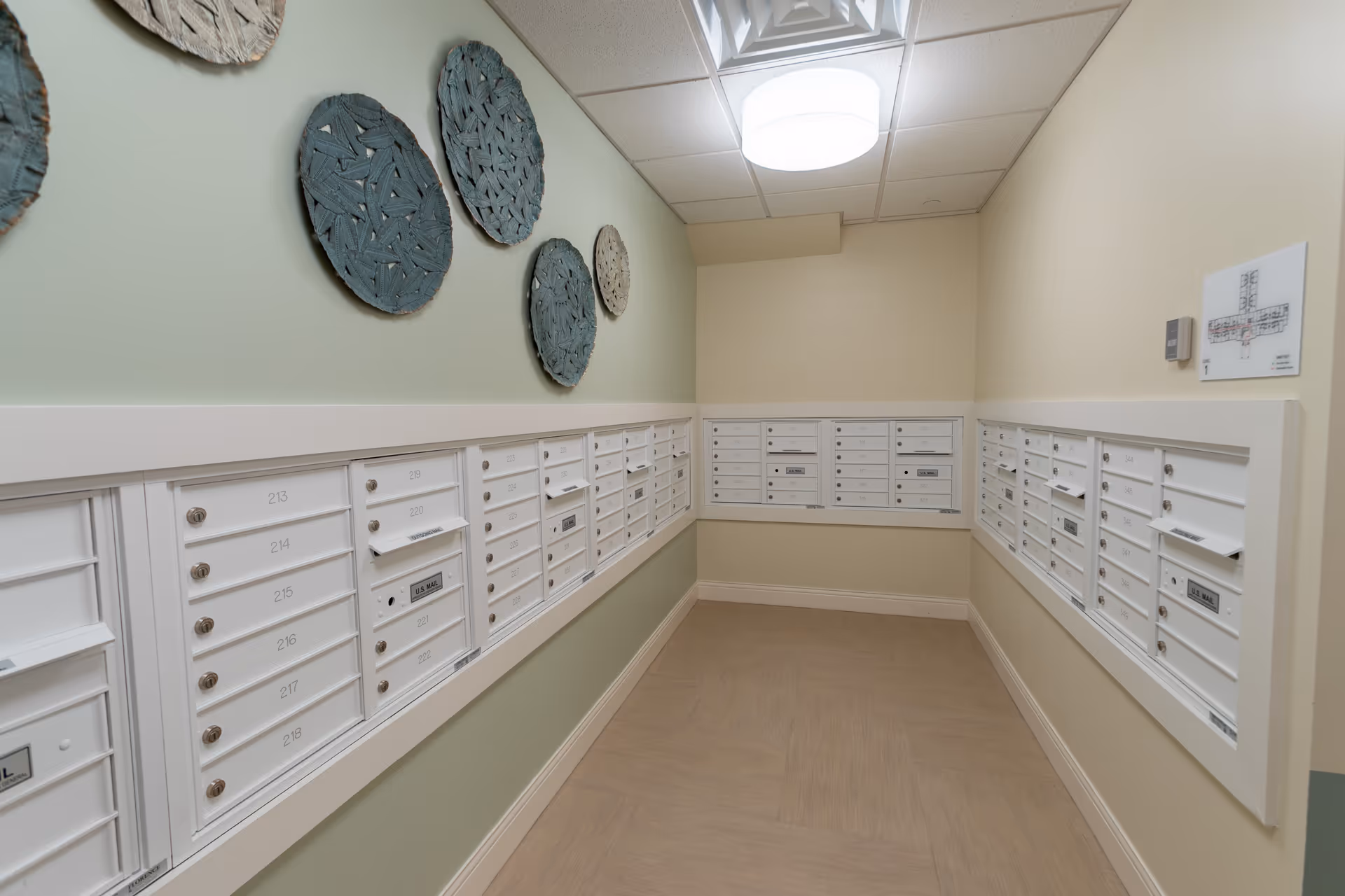 A mailroom area with multiple white mailboxes mounted on three walls. The walls are painted in light green and beige tones, and there are decorative woven circular wall hangings on the left wall. The floor has a light wood pattern, and a round ceiling light fixture illuminates the space. A floor plan is mounted on the right wall.