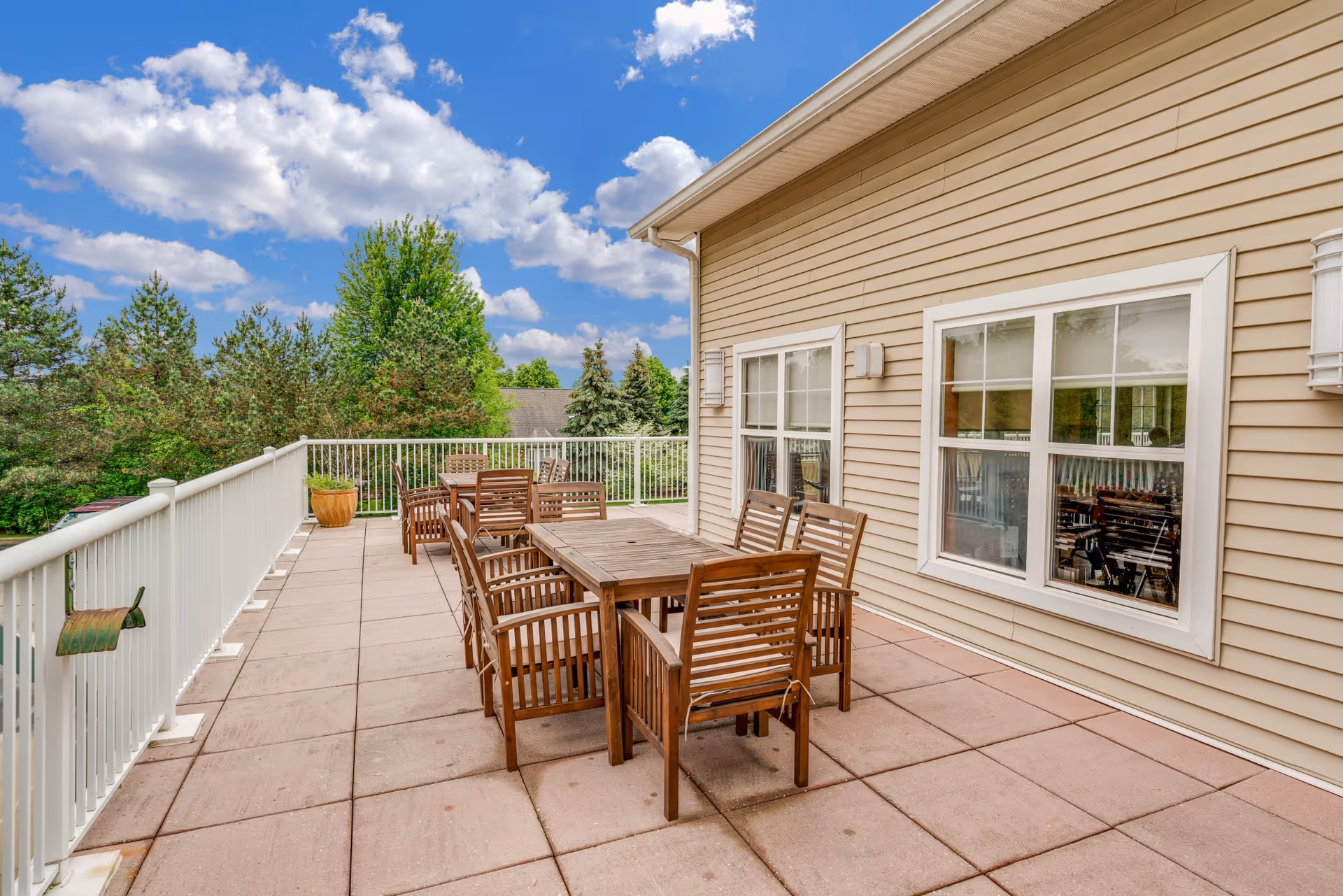 Outdoor patio area with wooden tables and chairs on a tiled floor next to a beige building with white-framed windows. The patio is surrounded by a white railing and overlooks green trees under a partly cloudy blue sky.