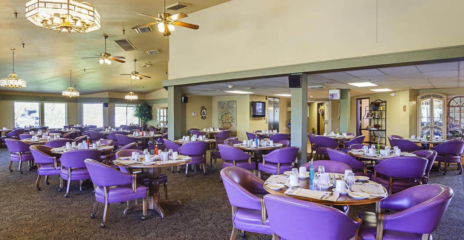 Spacious dining room with multiple round wooden tables set with plates, cups, utensils, and condiments. Each table is surrounded by purple cushioned chairs. The room has carpeted floors, ceiling fans, and large windows letting in natural light.