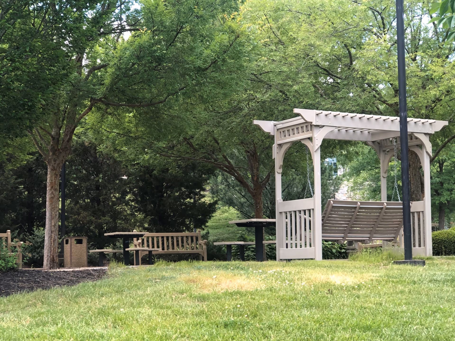 A peaceful outdoor garden area with green grass, trees, wooden benches, picnic tables, and a white wooden swing under a pergola structure.