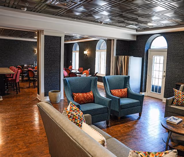 A cozy senior living common area with blue armchairs and a gray sofa adorned with colorful patterned pillows. The room features dark walls, arched windows with white shutters, a wooden floor, and a decorative ceiling. In the background, there is a dining area with tables and red chairs, and a door with glass panels leading outside.