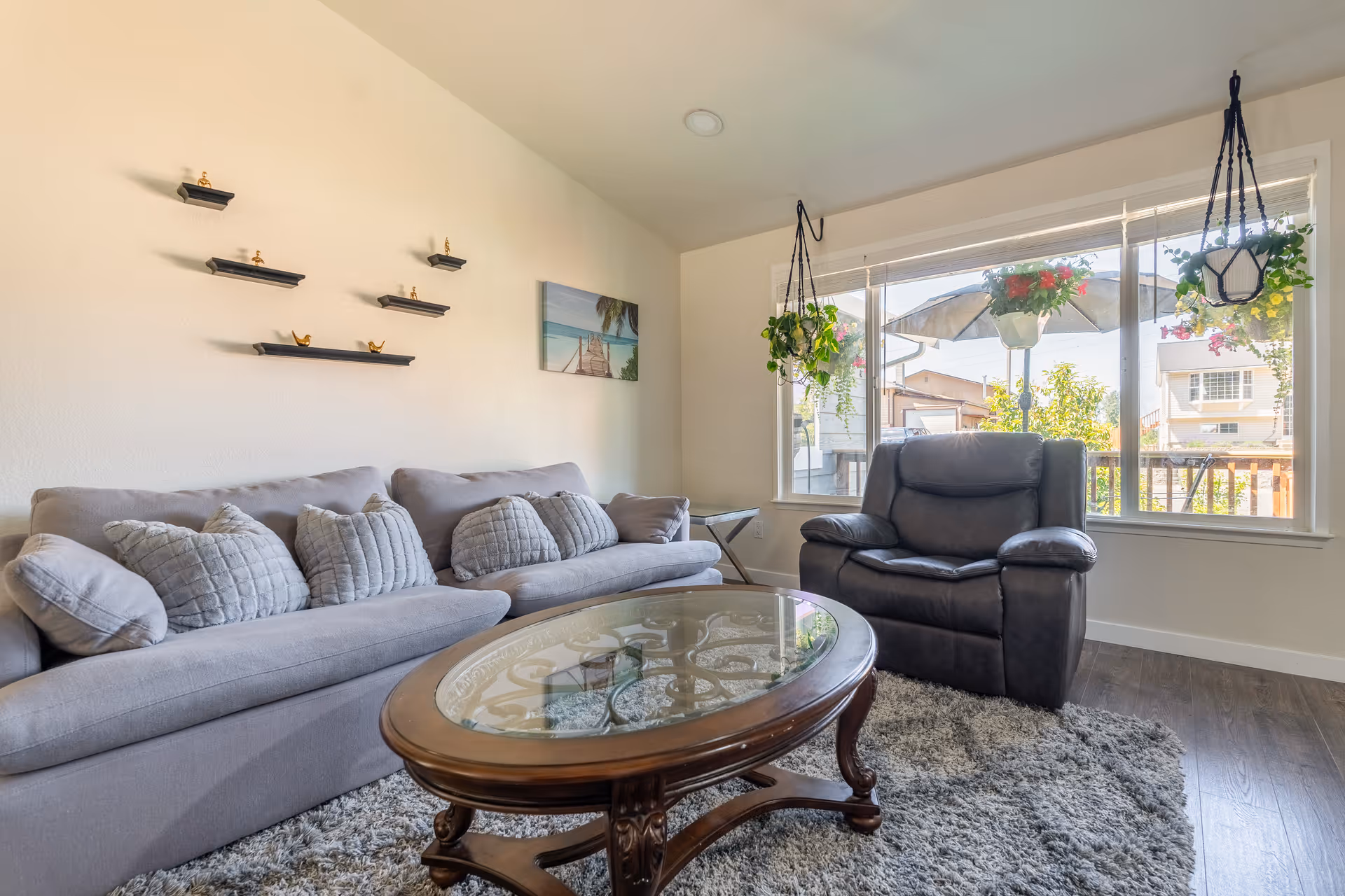 Bright living room with a gray sofa, leather recliner, glass-top coffee table and large window with hanging plants.