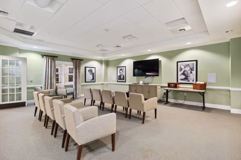 A well-lit room with light green walls and beige carpet featuring two rows of beige upholstered chairs facing a wall-mounted flat screen TV. The room has framed black and white portraits on the walls, a cabinet under the TV, a table with a basket and a radio, and a glass-paneled door with curtains on the windows.
