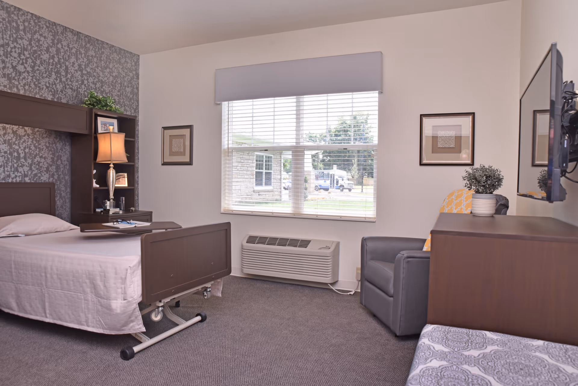 A senior living facility bedroom with a hospital-style bed, a bedside table with a lamp and framed photos, a window with blinds, a gray armchair with a yellow patterned pillow, a wooden dresser with a potted plant, and a wall-mounted TV.
