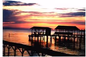 A serene sunset over a body of water with two covered wooden docks extending into the water, silhouetted against the colorful sky with shades of orange, pink, and purple.