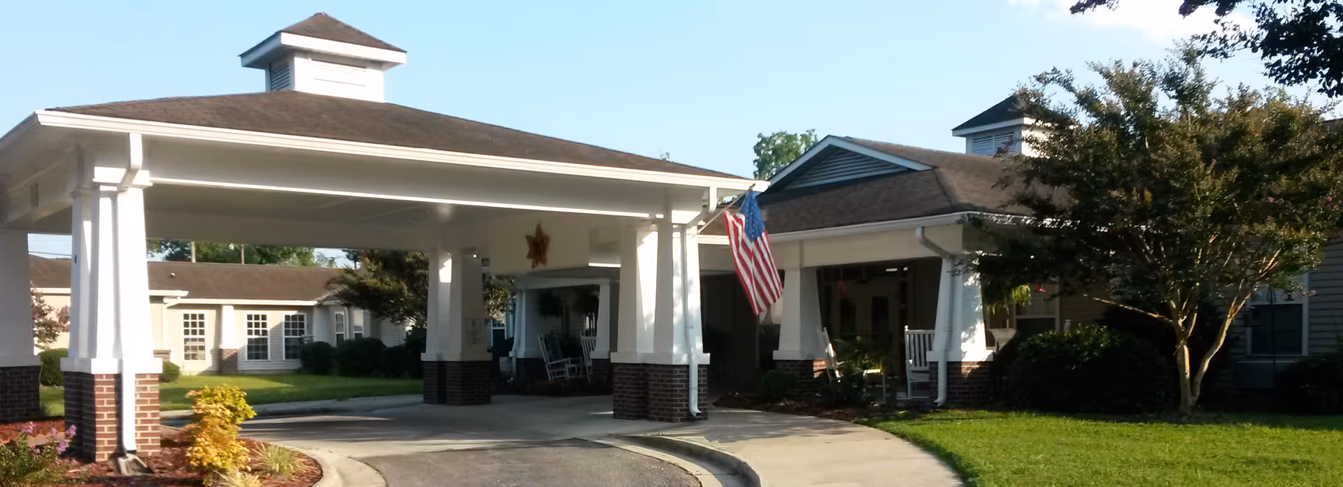 Entrance of Somerset Court of Rocky Mount senior living facility with a covered drop-off area, white columns with brick bases, an American flag, rocking chairs on the porch, and well-maintained landscaping under a clear blue sky.