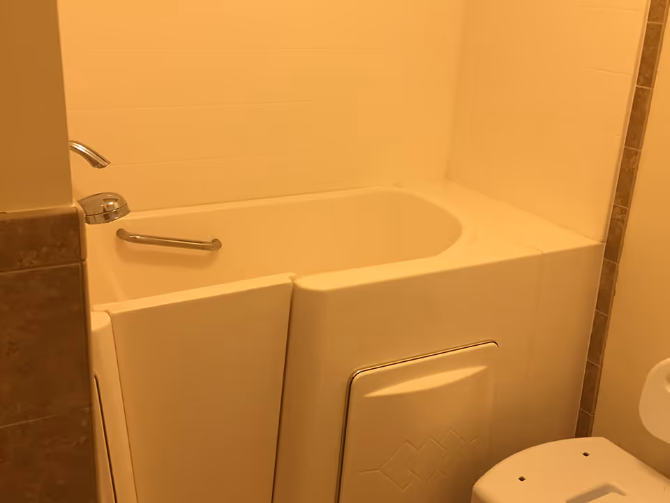 A walk-in bathtub with a grab bar and faucet in a bathroom, with part of a toilet visible at the right.