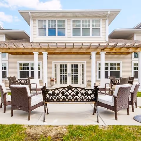 Outdoor patio with wicker chairs, a decorative metal bench and pergola in front of a two-story building.