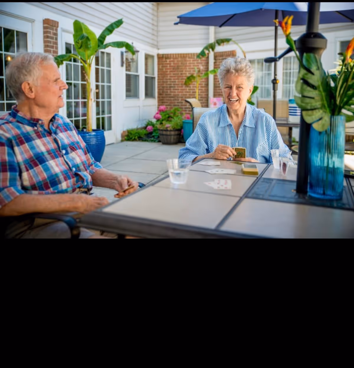 Two elderly individuals sitting at an outdoor table playing cards. The setting is a patio area with potted plants, a large umbrella, and a building with windows and brick walls in the background. One person is wearing a blue plaid shirt and the other is wearing a blue and white striped shirt, smiling while holding cards.
