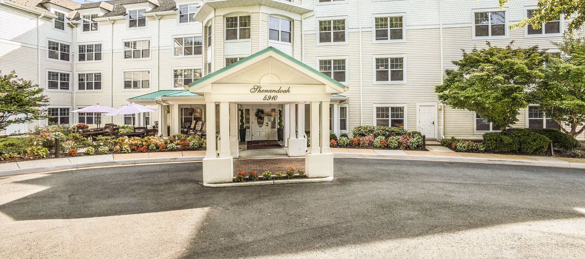 Front exterior view of a multi-story senior living facility named Shenandoah with white siding, multiple windows, a covered entrance with the number 5910, and landscaped flower beds and trees around the driveway.