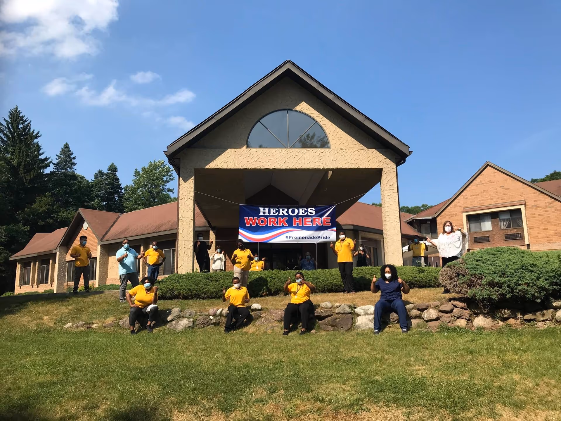 Staff wearing masks stand and sit on a lawn in front of a senior living building under a banner that reads 'HEROES WORK HERE'.