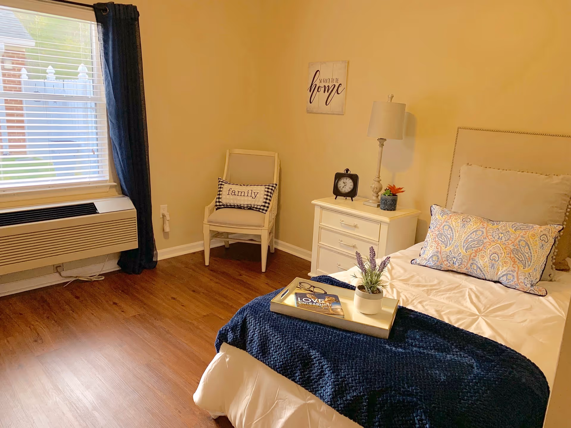 A cozy bedroom with a single bed featuring a beige headboard, white bedding, a blue knitted throw blanket, and a decorative pillow with a paisley pattern. A white nightstand beside the bed holds a table lamp, a small clock, and a potted plant. A chair with a cushion labeled 'family' is placed near a window with blinds and dark curtains. A wall decoration above the nightstand reads 'So good to be home'. The room has wooden flooring and beige walls.