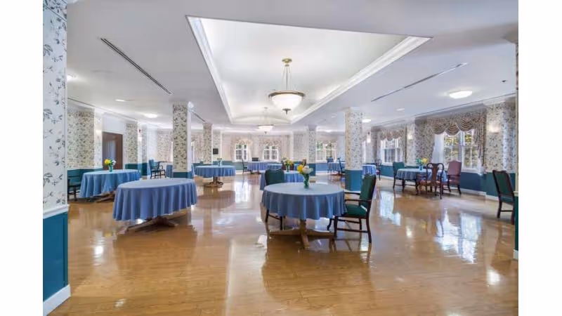Spacious dining room with multiple round tables covered with blue tablecloths, each table having a small flower arrangement. The room has wooden floors, floral wallpaper, several windows with draped curtains, and ceiling light fixtures.