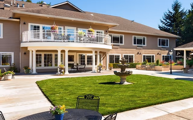 Outdoor courtyard area of The Hampton and Ashley Inn Memory Care Community featuring a well-maintained green lawn with a tiered water fountain in the center, surrounded by paved walkways. The two-story building has a balcony with hanging flower pots and several windows. There are outdoor tables and chairs for seating, along with potted plants and a lamppost.