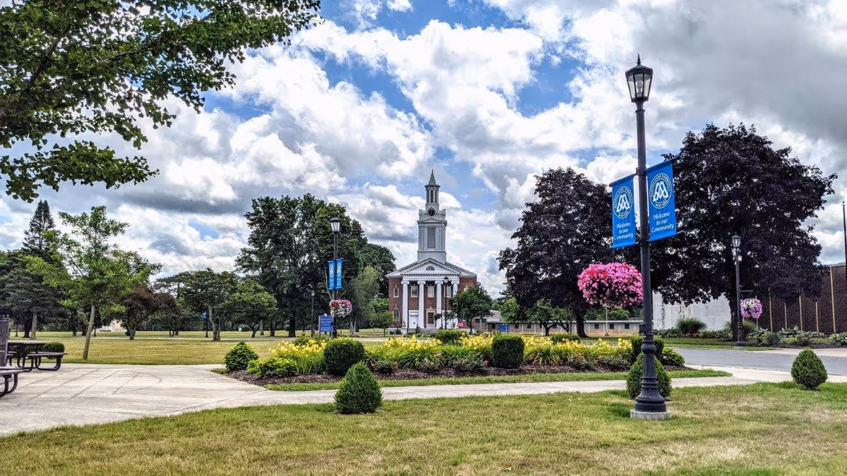 Outdoor view of the Masonic Care Community of New York featuring a well-maintained garden with green grass, shrubs, and flowers. There are several trees and lamp posts with hanging flower baskets and blue banners that say 'Welcome to our Community.' In the background, a large building with a white clock tower and columns is visible under a partly cloudy sky.