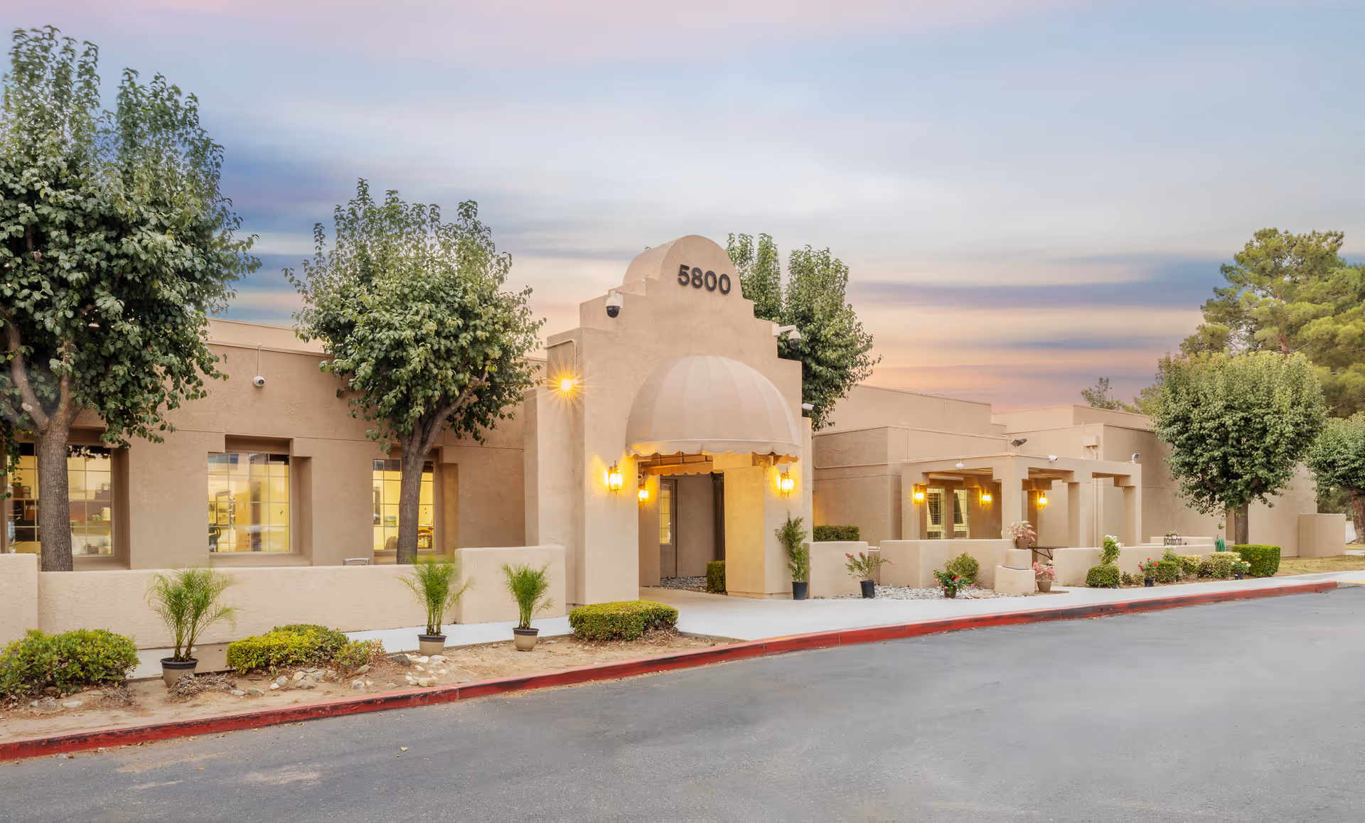 Front exterior of the Sundance Creek Post Acute building with an arched entrance marked "5800", trees, and low landscaping at dusk.