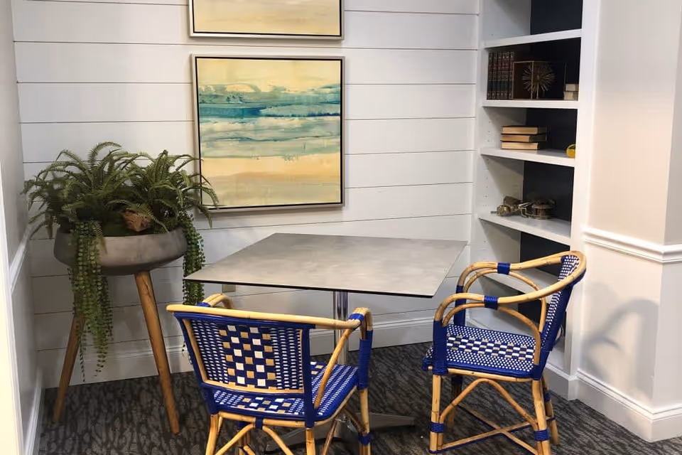 A small corner seating area with a square table and two blue and beige woven chairs. There is a tall plant in a pot on wooden legs to the left and a built-in white bookshelf with books and decorative items to the right. Two abstract paintings with blue and beige tones hang on the white shiplap wall behind the table.