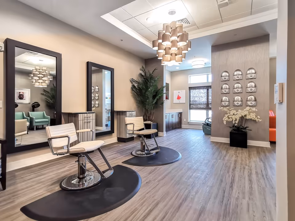 Bright salon-style common area with two styling chairs, large mirrors, decorative lighting, and seating in a senior living facility.