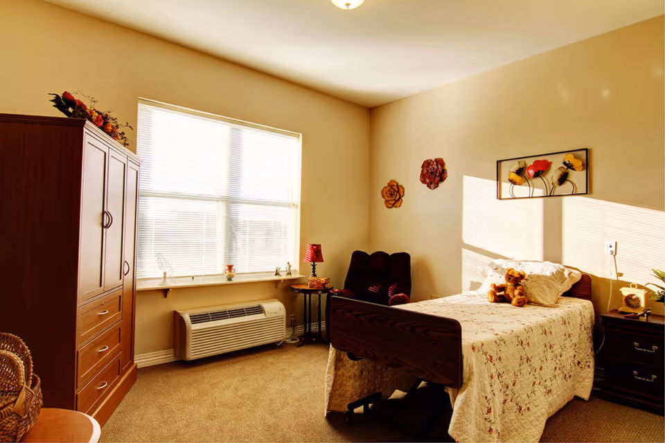 A cozy bedroom with a single bed covered in a floral quilt and a teddy bear on top. There is a dark wooden nightstand with a clock and a small plant, a burgundy armchair, and a wooden wardrobe. The room has beige walls decorated with flower wall art and a large window with blinds letting in sunlight.