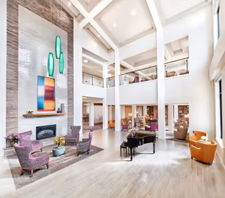 Bright and spacious two-story living room area with high ceilings, large windows, and modern decor. The room features a fireplace with colorful artwork above it, three purple armchairs arranged around a small table with flowers, a black grand piano, and additional seating including orange chairs near the windows. The upper level has a balcony with glass railings overlooking the space.