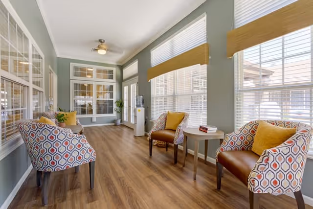 A bright and airy sitting area with large windows covered by white blinds and yellow valances. The room features wood flooring, a ceiling fan, and four patterned armchairs with yellow cushions arranged around two small round tables. A water cooler is placed near the far wall, and there are large interior windows on the left side.