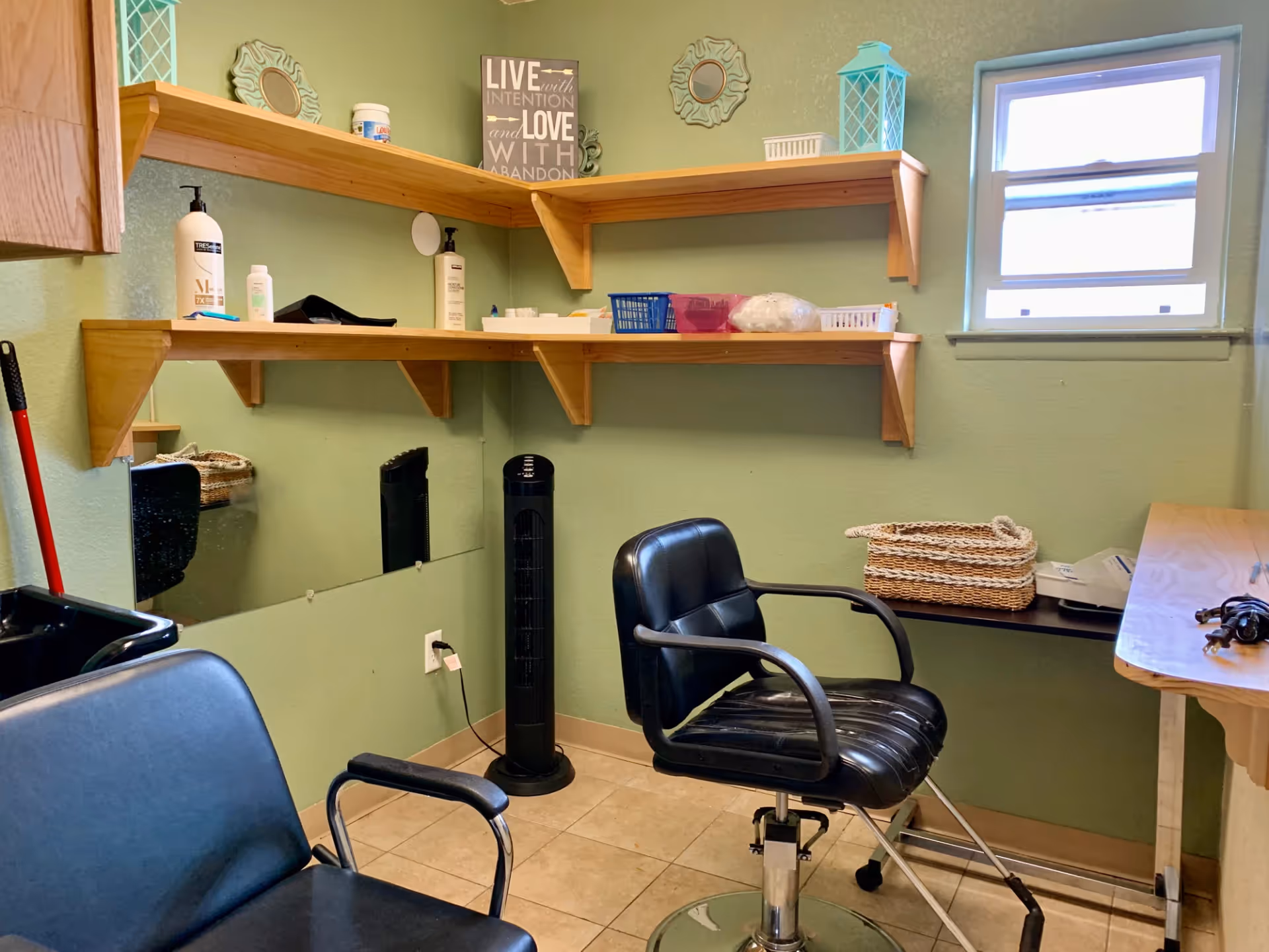 A small room with green walls featuring two black chairs, one resembling a salon chair with wheels and adjustable height. There are wooden shelves mounted on the walls holding various items including bottles, a basket, and decorative pieces. A small window lets in natural light, and a tall black fan stands against the wall near a mirror.