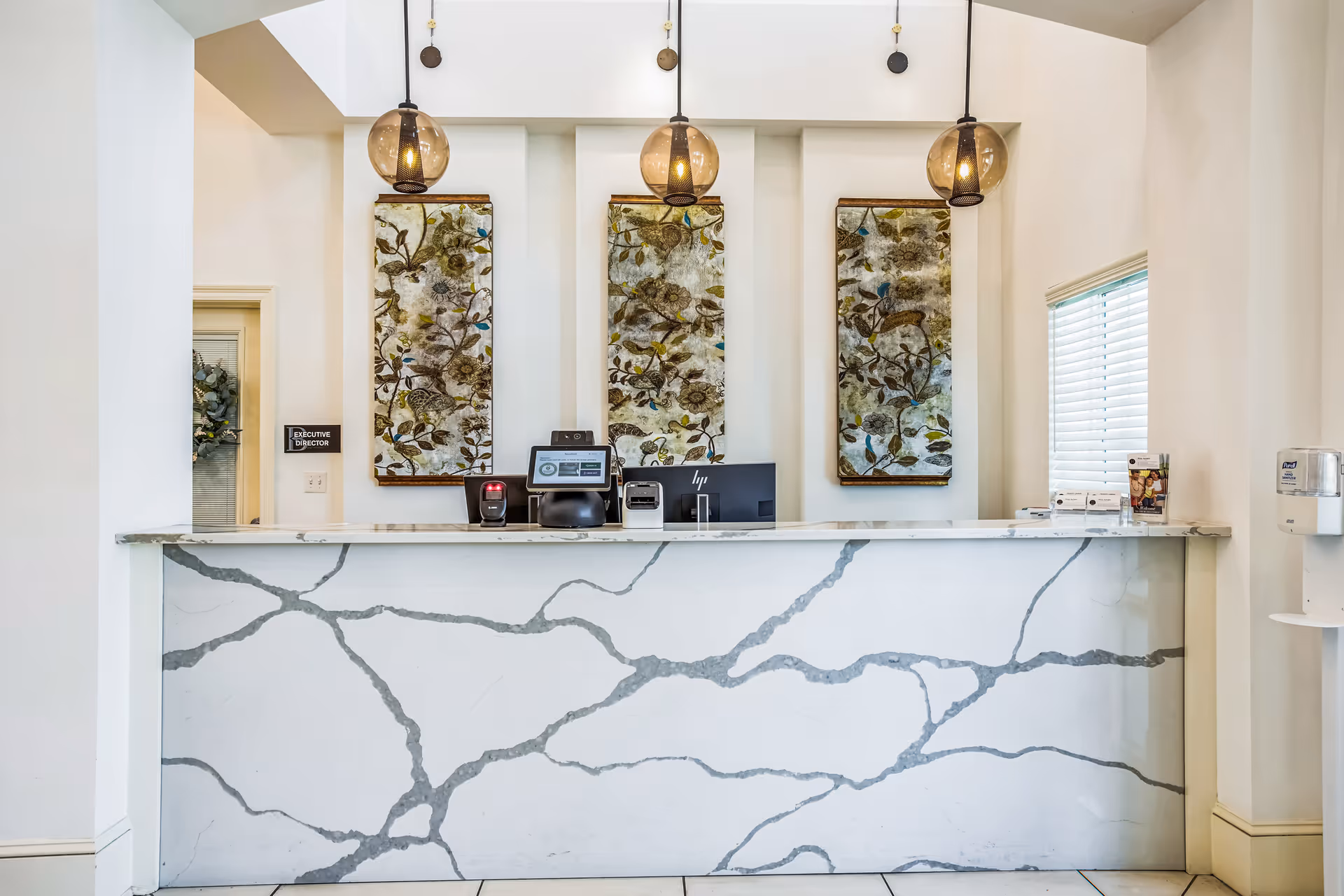 Reception desk area with a white marble counter featuring gray veining, three pendant lights hanging from the ceiling, and three vertical floral artworks on the wall behind the desk. There is a computer monitor, a printer, and other office equipment on the desk. A sign on the left wall reads 'Executive Director'.