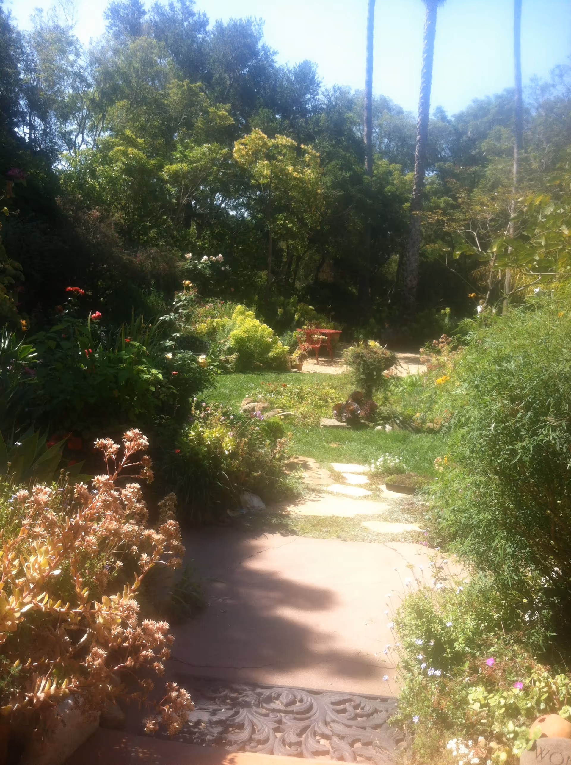 A lush garden path surrounded by various green plants, bushes, and flowering plants leading to a small red table and chairs set in the distance, with tall trees and a clear blue sky overhead.