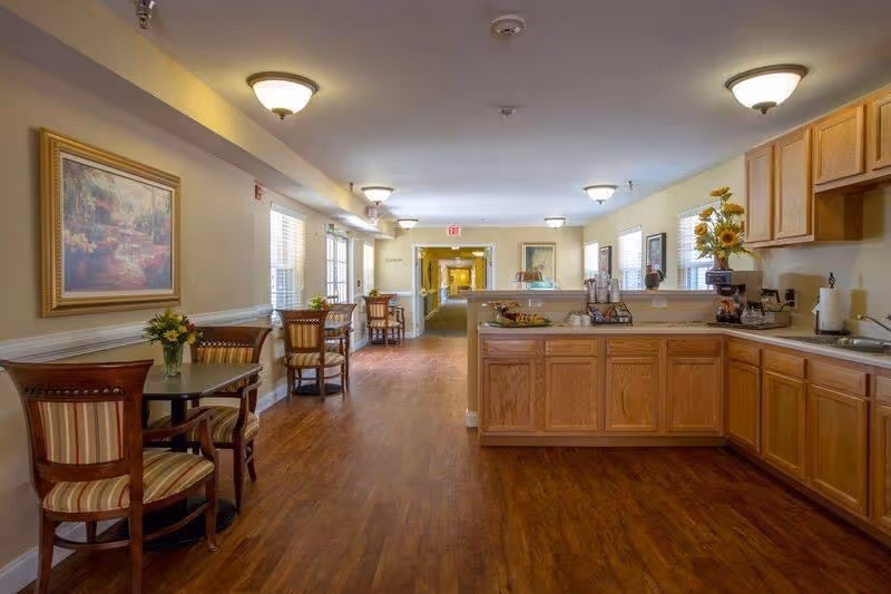 A bright communal dining area with round tables and chairs, wood flooring, and a kitchenette with oak cabinets on the right.