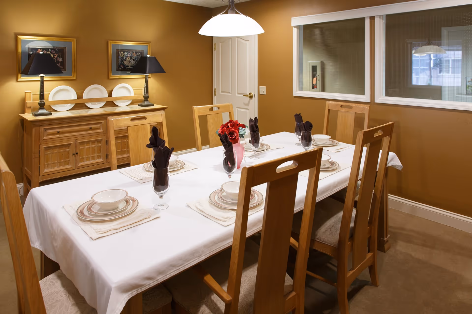A dining room with a rectangular table covered with a white tablecloth, set with plates, bowls, glasses with dark napkins, and a pink vase with red flowers in the center. The room has six wooden chairs around the table, a wooden sideboard with two lamps and decorative plates, two framed pictures on the wall, and a hanging ceiling light above the table. The walls are painted brown, and there are two windows with blinds on one side.
