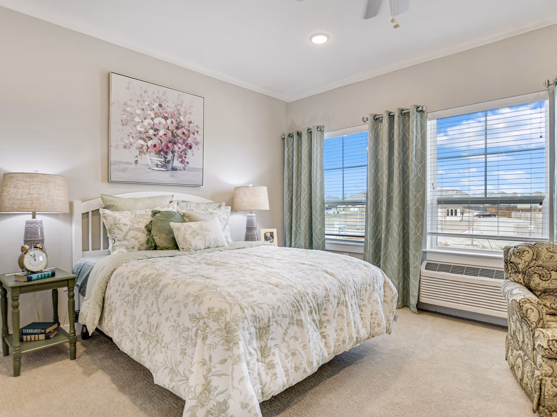 A bright bedroom with a large bed covered in floral patterned bedding and multiple pillows. There are two bedside tables each with a lamp, an alarm clock, and books. A floral painting hangs above the bed. The room has two large windows with green patterned curtains, letting in natural light and showing a view of a parking lot outside. A patterned armchair is partially visible on the right side.