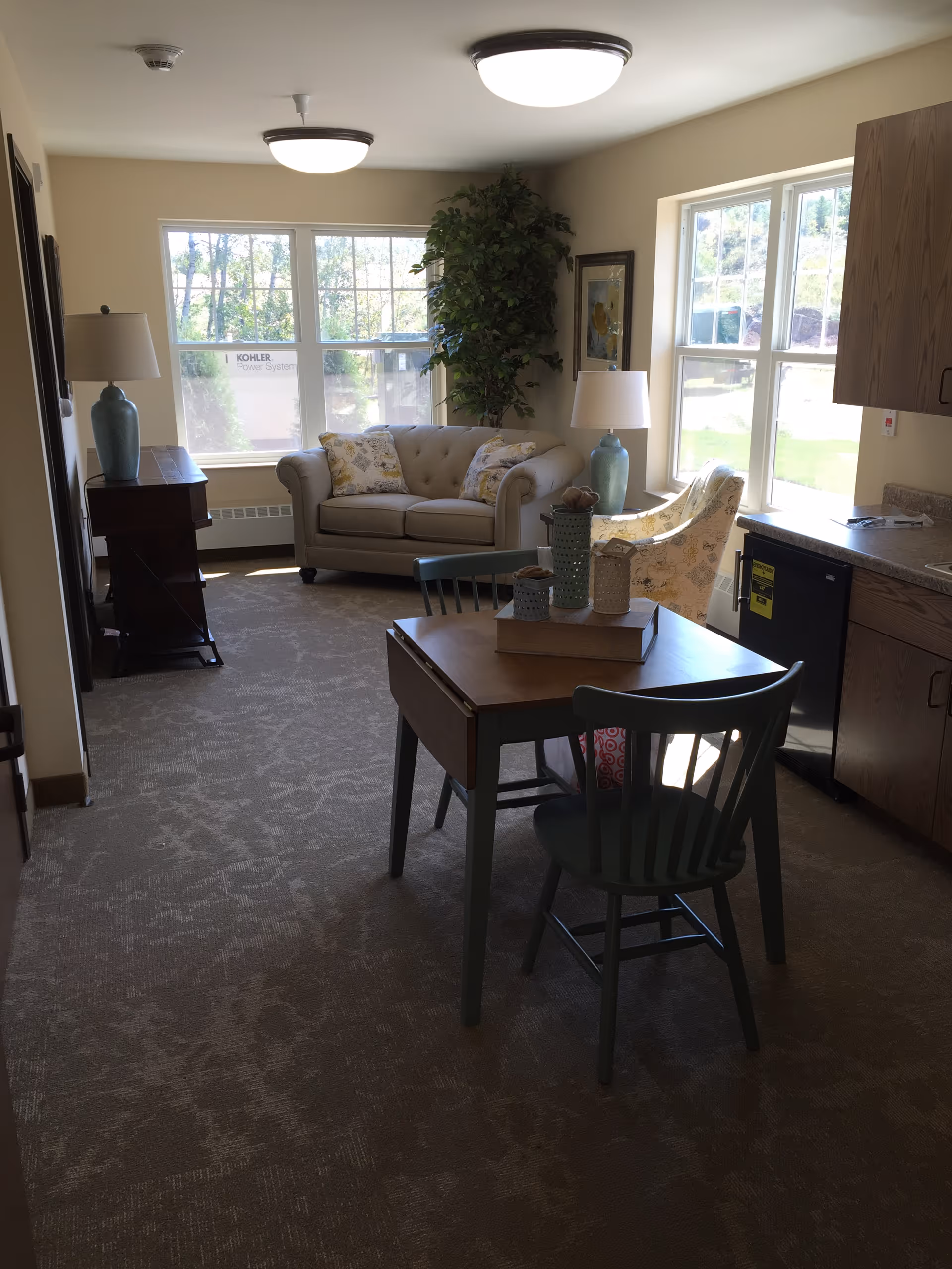 A cozy senior living community room with a small wooden dining table and two chairs in the foreground. In the background, there is a beige loveseat with floral pillows, a floral armchair, a tall green plant, and two table lamps with blue bases. Large windows allow natural light to fill the room, and there is a small kitchenette area with wooden cabinets and a mini fridge on the right side.