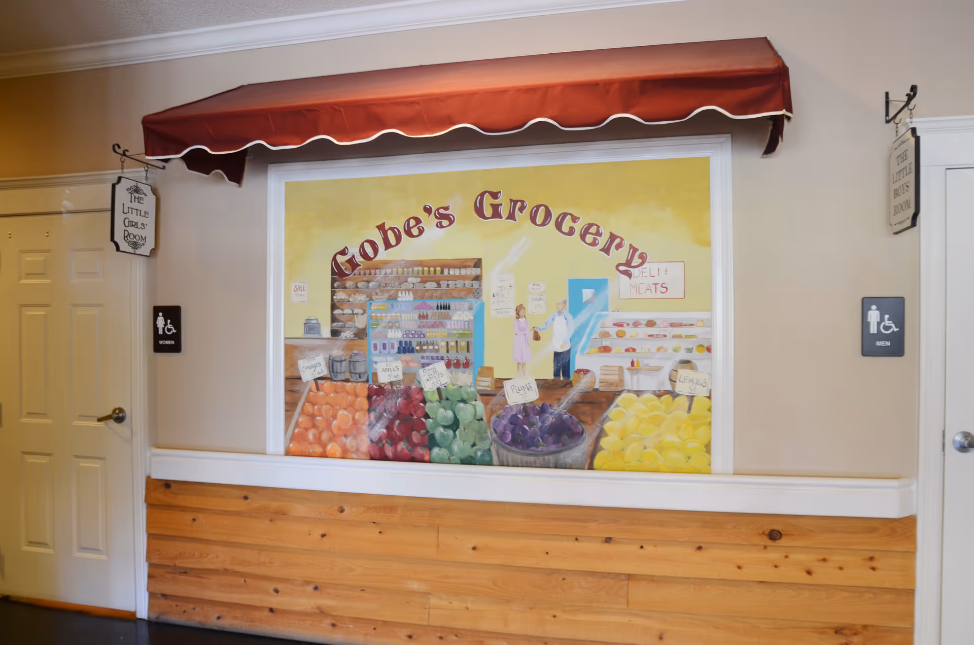Interior hallway wall featuring a painted 'Gobe's Grocery' mural under a red awning between doors marked men's and women's restrooms.