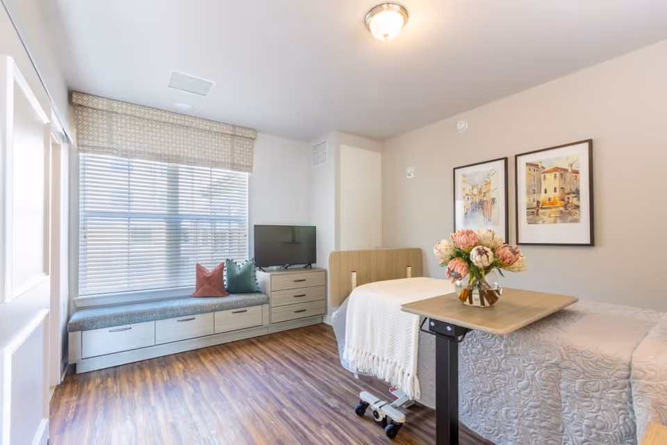 A bright and clean bedroom in a senior living facility featuring a single bed with a white quilt and a wooden headboard. A small overbed table with a vase of flowers is positioned over the bed. There is a window seat with cushions and storage drawers beneath, next to a small dresser with a flat-screen TV on top. Two framed paintings hang on the beige wall above the bed. The room has wood flooring and a ceiling light fixture.