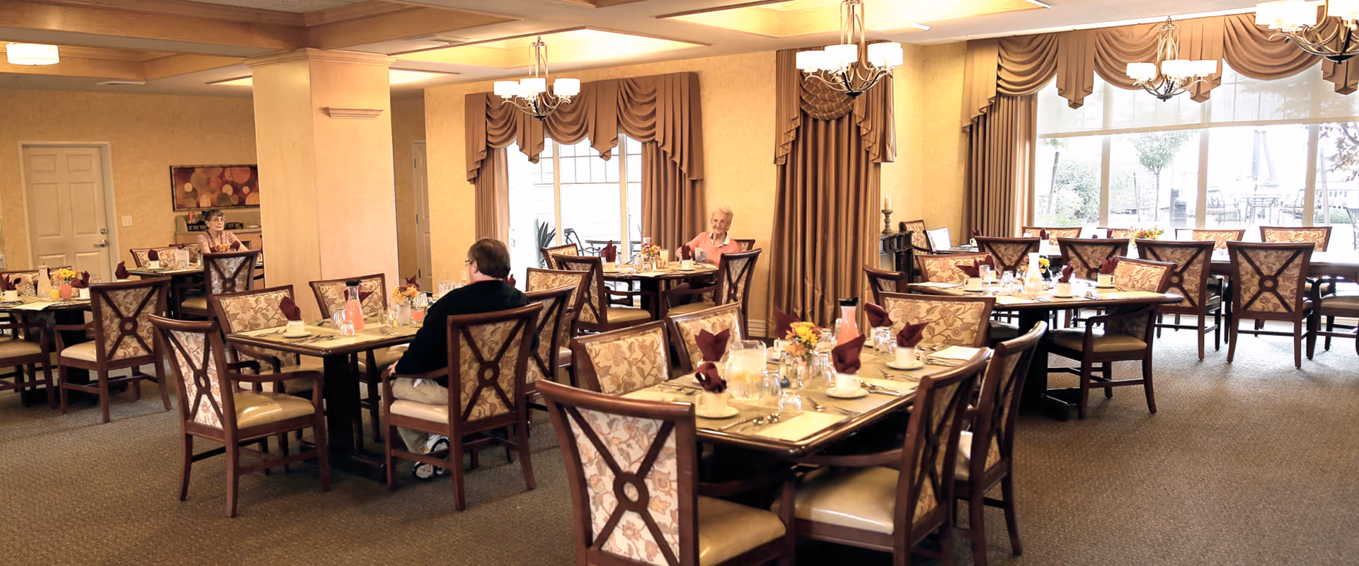 A spacious dining room in a senior living facility with multiple tables set with plates, glasses, and napkins. Several elderly residents are seated at different tables. The room has large windows with curtains allowing natural light to enter, and chandeliers hanging from the ceiling provide additional lighting.