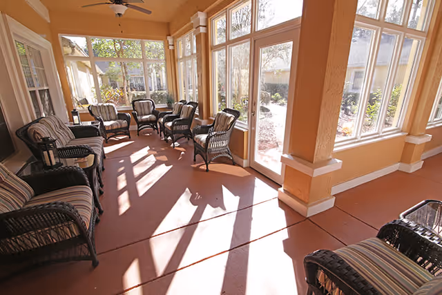 Sunlit enclosed sunroom with wicker chairs and small tables arranged along large windows.