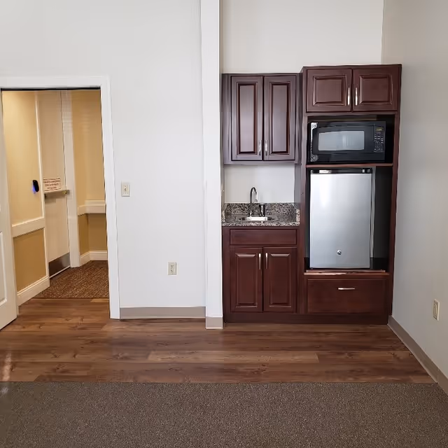 Interior view of a senior living facility room showing a small kitchenette area with dark wood cabinets, a granite countertop with a sink, a microwave, and a mini refrigerator. The room has wood flooring near the kitchenette and carpeted flooring in the foreground. There is a doorway leading to a hallway with beige walls and carpet.