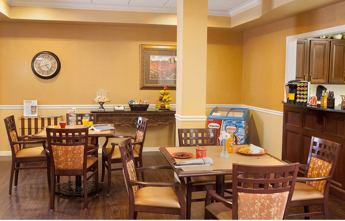 A cozy dining area with wooden tables and chairs featuring patterned cushions. On the tables are plates with food, drinks, a newspaper, and a flower vase. The room has warm yellow walls, a clock, a framed picture, and a sideboard with snacks and a fruit stand. There is also a small freezer with ice cream branding and a coffee station with various beverages.