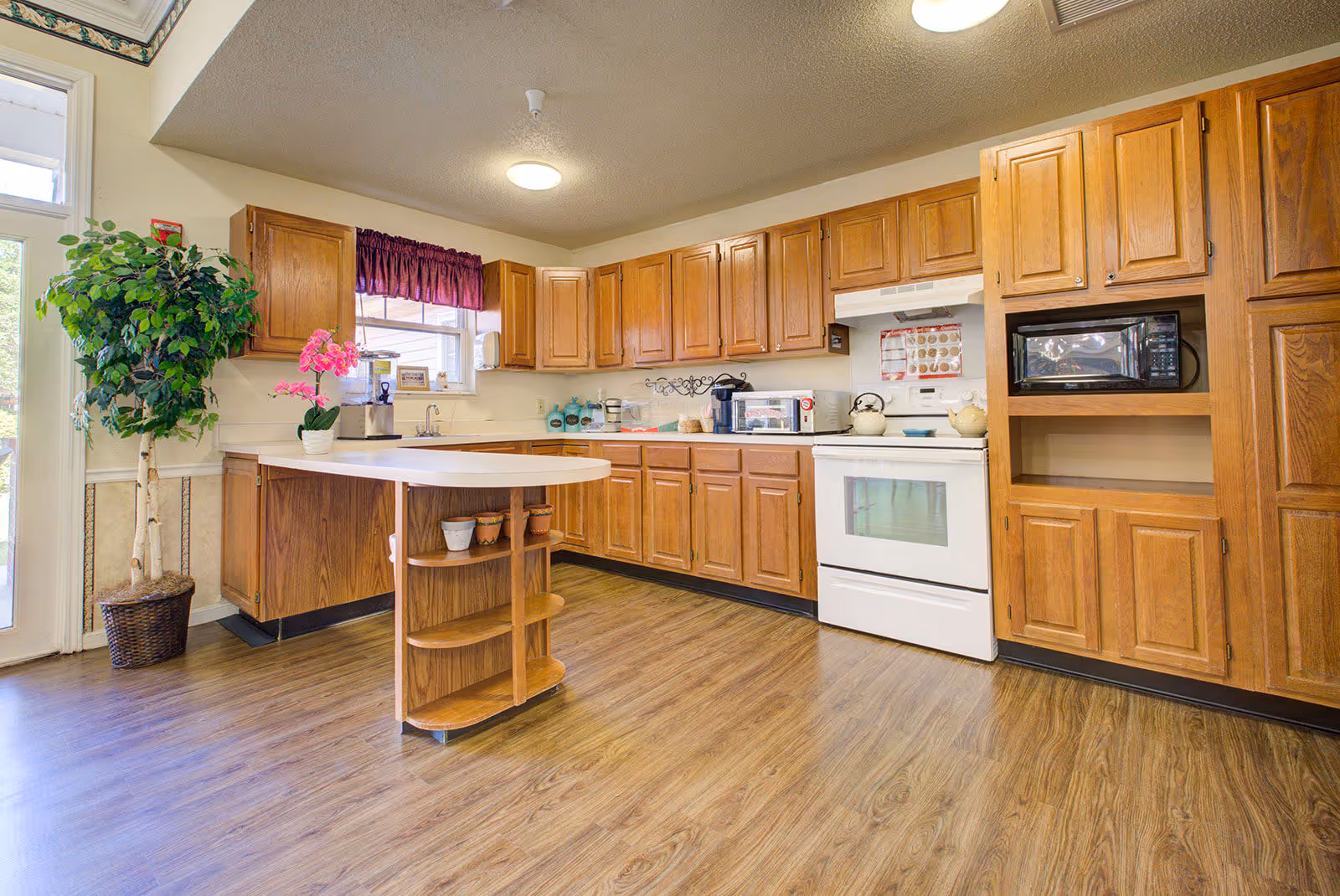 Bright kitchen with wooden cabinets, a white stove, microwave, and a small island with shelves. There is a potted plant and a pink orchid on the counter near a window with a purple valance.
