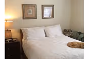 A bedroom with a neatly made bed featuring white bedding and pillows. There are two framed pictures hanging on the wall above the bed. A bedside table with a lamp and some items is positioned to the left of the bed. A cat is lying on the right side of the bed.
