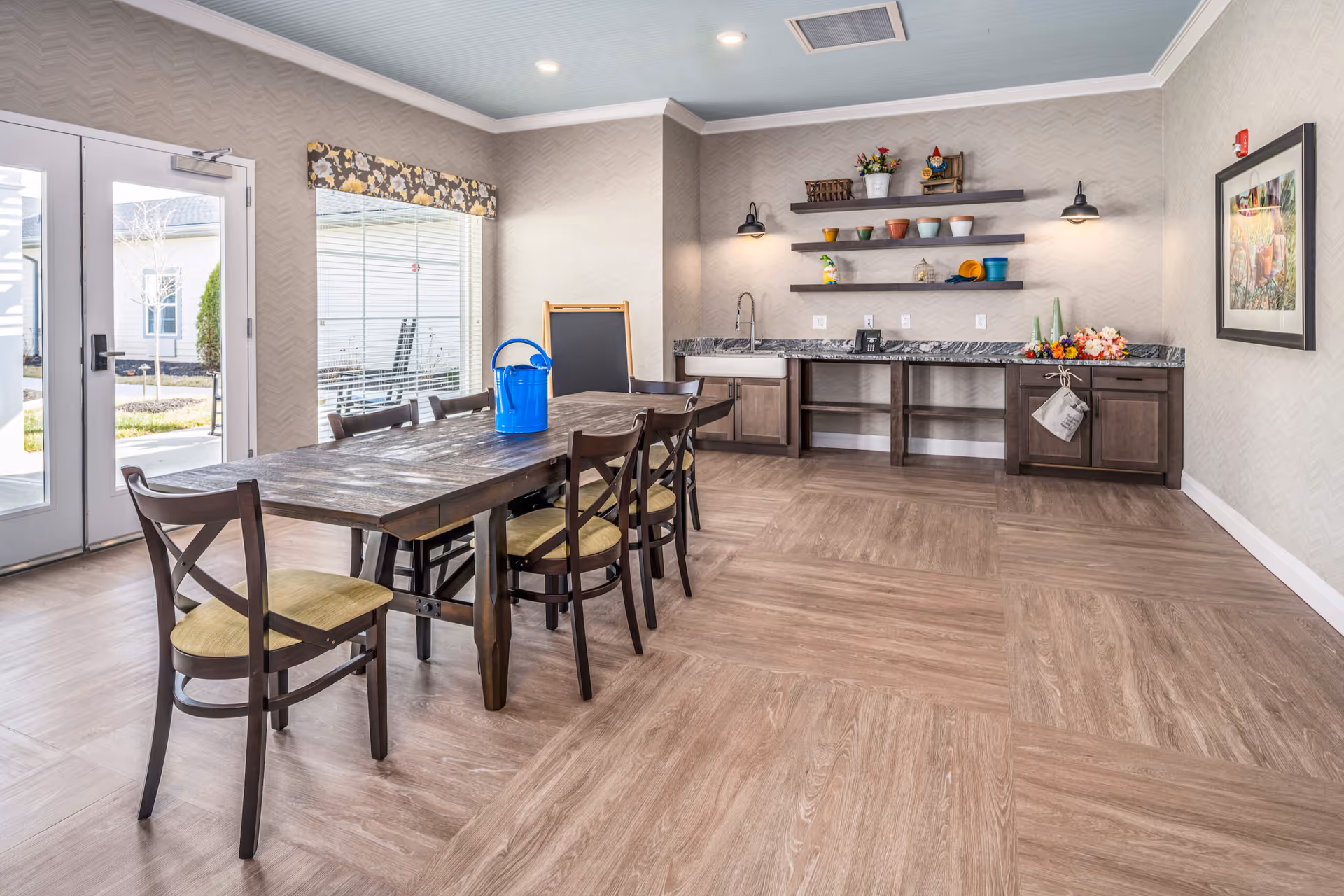 Bright communal dining room with a long wooden table and chairs, a countertop with sink and open shelves, and double glass doors to the outside.