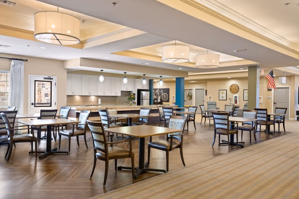 Bright communal dining room with multiple square tables and chairs, a serving counter and pendant lighting.