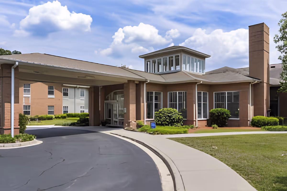 Exterior view of TerraBella Windsor Lake senior living facility showing the building entrance with a covered driveway, large windows, and surrounding greenery under a partly cloudy sky.