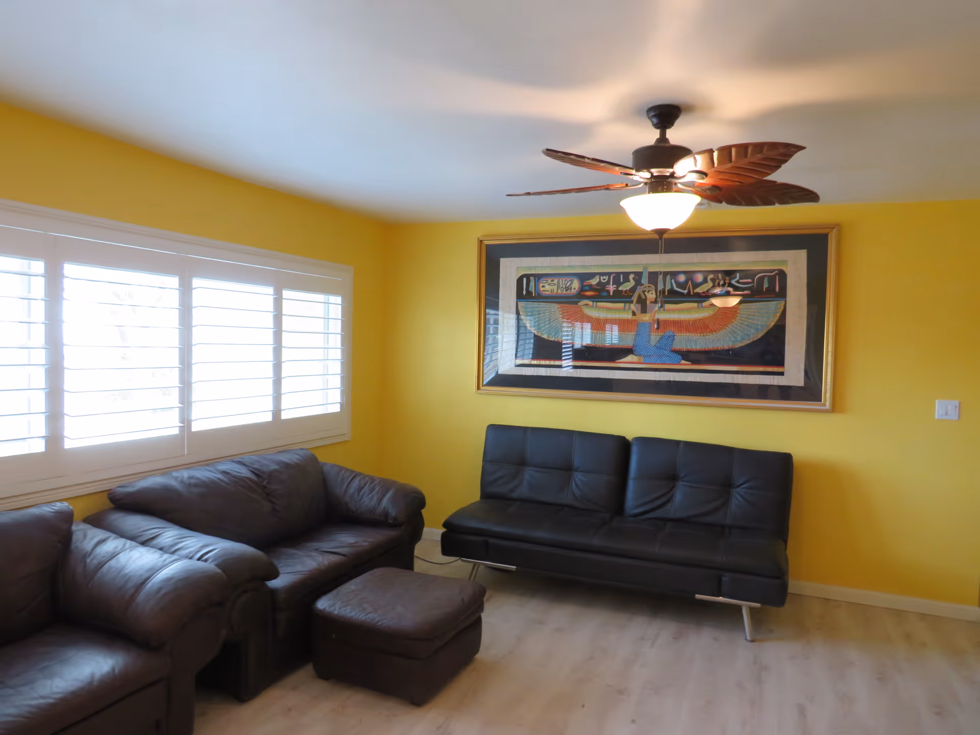 A living room with yellow walls featuring a large window with white shutters on the left. The room contains a dark brown leather armchair, a matching loveseat, a dark brown leather ottoman, and a black leather futon. A ceiling fan with leaf-shaped blades and a light is mounted on the ceiling. A large framed artwork with an Egyptian theme hangs on the wall above the futon.